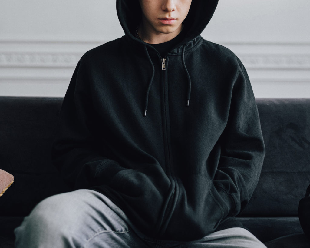 Psihološka podpora - resen mladostnik v črni jopici s kapuco poveznjeno čez glavo in rokami v žepih